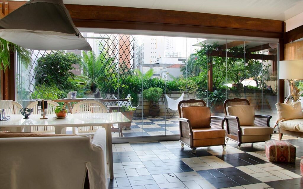 ambiente agradável com mesa, cadeiras e poltronas, envidraçado com vista para varanda externa repleta de plantas