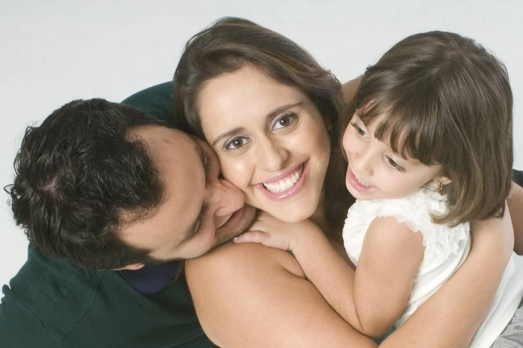retrato familiar, mãe no meio, olhando para a lente da câmera, com filha no colo do lado direito e pai á esquerda olhando para a criança