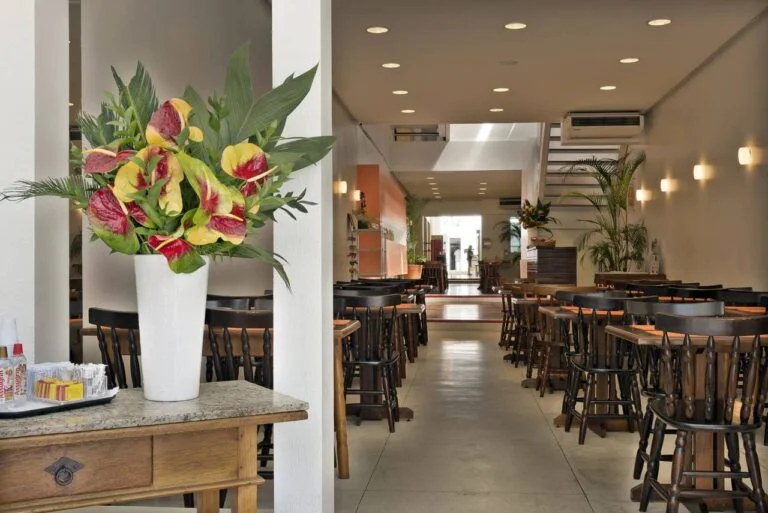 ambiente de restaurante, com luz suave,mesas e cadeiras de madeira, organizadas e postas com jogos americanos na cor laranja, em primeiro plano uma bancada pequena com adoçantes e açucares organizados ao lado de um vaso com arranjo de plantas com folhas variadas nas cores verde, amarelo e vermelho