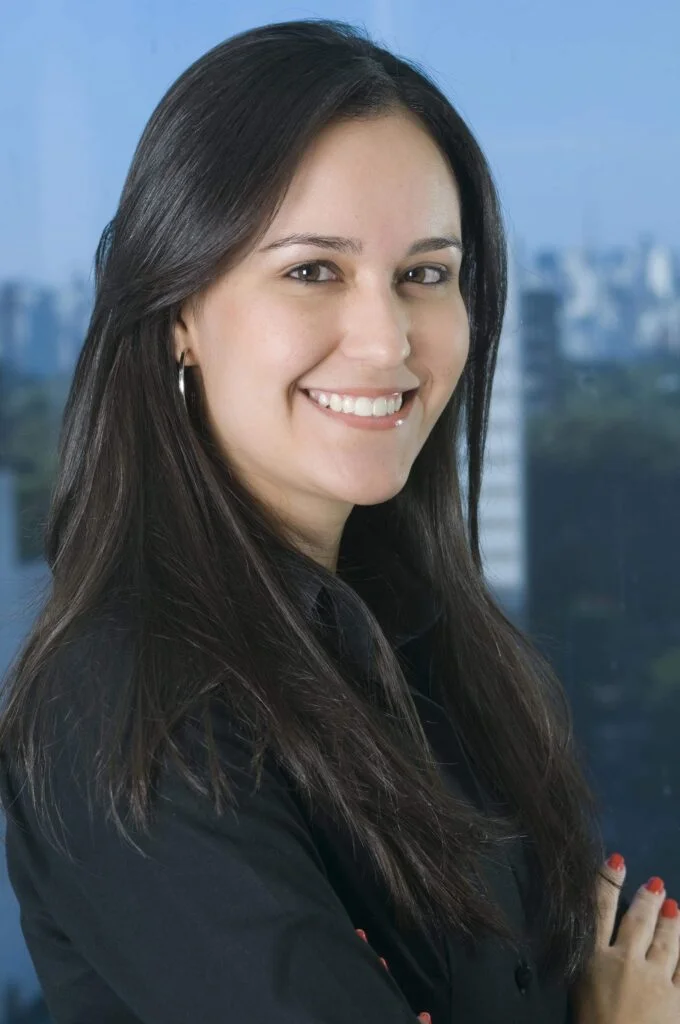 retrato profissional de mulher jovem branca, cabelos castanhos e lisos, sorrindo, á frente de uma janela