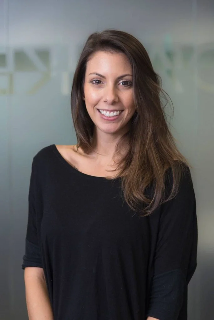 retrato profissional de mulher jovem de cabelos castanhos abaixo dos ombros, sorrindo, ambiente de escritório com fundo cinza desfocado