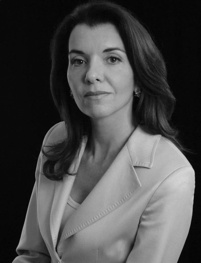 retrato profissional de mulher, pele clara e cabelos escuros pouco abaixo dos ombros, vestida com blazer claro, ambiente de fundo escuro, foto em preto e branco