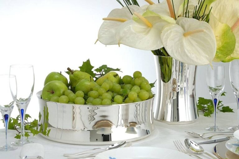 mesa posta com objetos de prata, travessa com uvas verdes, jarra de prata com arranjo de antúrios brancos, taças de vidro e talheres