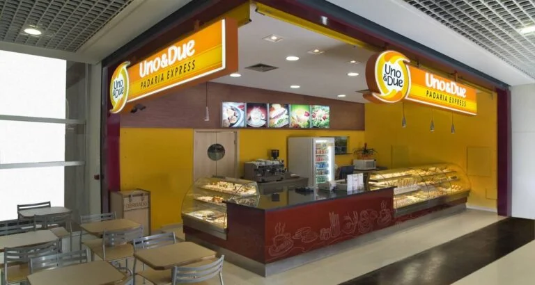 Quiosque Uno&Due, com os letreiros e vitrines acesos, vitrine bem completa de opções doces e salgadas, algumas mesas para realizar o consumo ao lado esquerdo, todo o ambiente limpo e organizado, sem pessoas