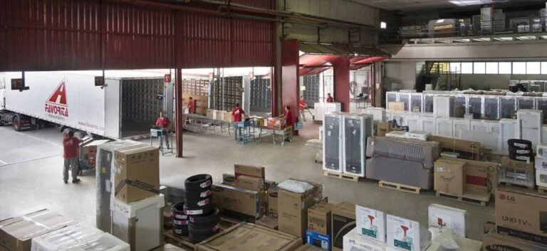 ambiente de deposito, contendo muitos eletrodomésticos, móveis, dentre outras coisas, com funcionários em função de carregamento de caminhões