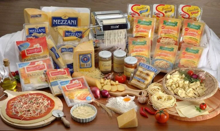 mesa de madeira com uma variedade de massas cruas embaladas, á frente destas algumas opções de massa cruas em cesto com temperos, uma máquina á manivela de espagueti da marca Shule, algumas opções de queijos, uma pizza montada sobre tábua própria, além de cebolas rochas, tomates, pimentas, alho, farinha de trigo, ovo e jarro de azeite dispostos aleatoriamente sobre a mesa