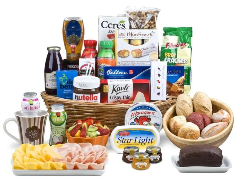 cesta de café da manhã recheada com guloseimas, bebidas lácteas, sucos, biscoitos, mel e creme de avelã, rodeada por frios fatiados, iogurtes, pães, bolo, geléias, manteigas e frutas e, base branca