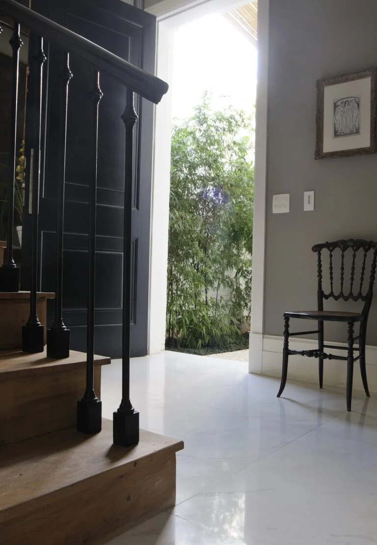 hall de entrada, mostra primeiros degrais de escada em madeira com corrimão preto, piso semelhante a mármore branco, parede cinza claro, com pequeno quadro, cadeira de madeira ornamental ao lado da porta aberta ,mostrando canteiro bamboos
