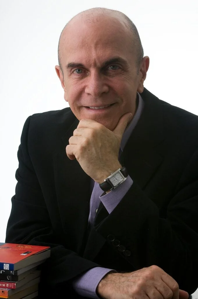 retrato profissional de homem de terceira idade,vestido com terno preto e camisa violeta, com mão sob o queixo, alguns livros na sua lateral