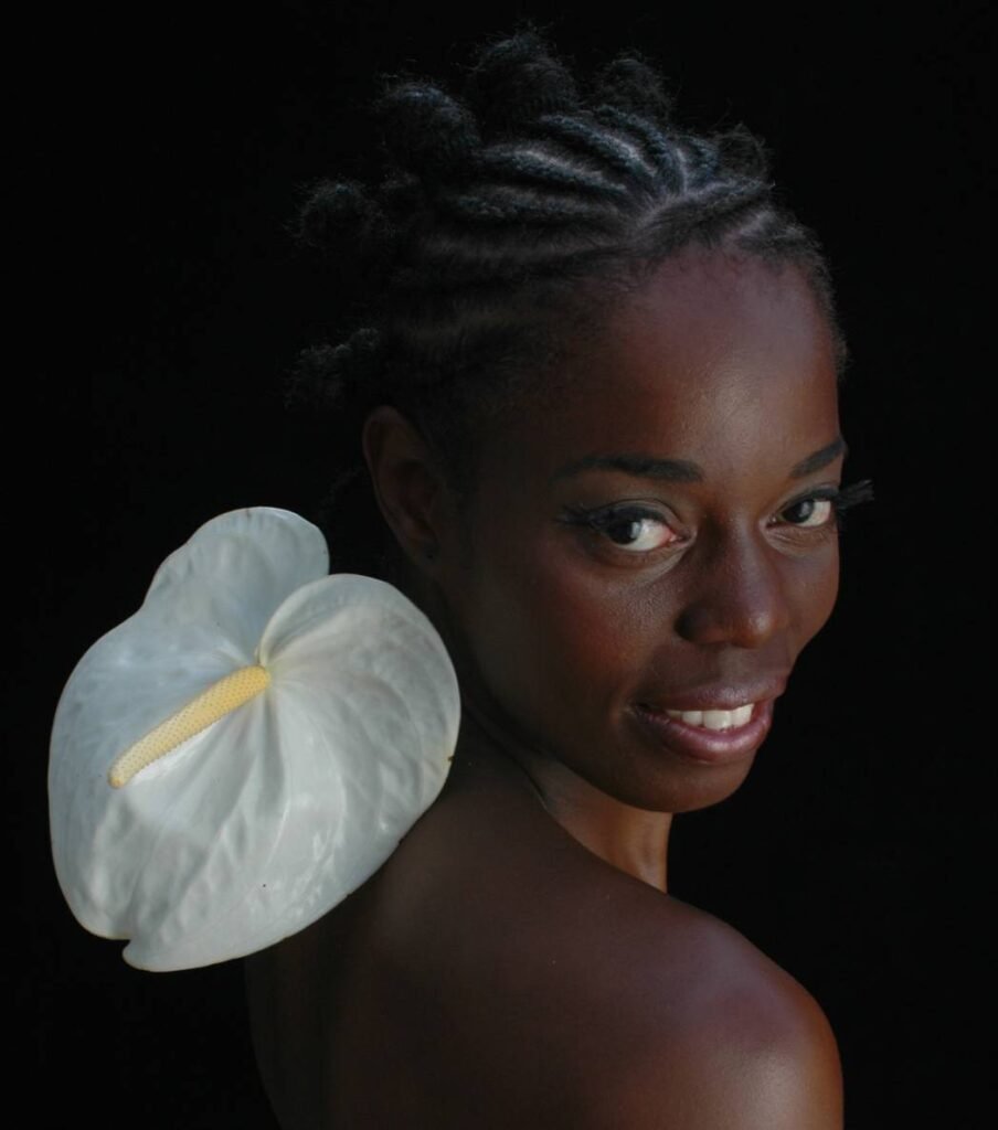 mulher negra, sorrindo, de costas com ombro e rosto direcionados para a câmera, ombros nus com flor de antúrio