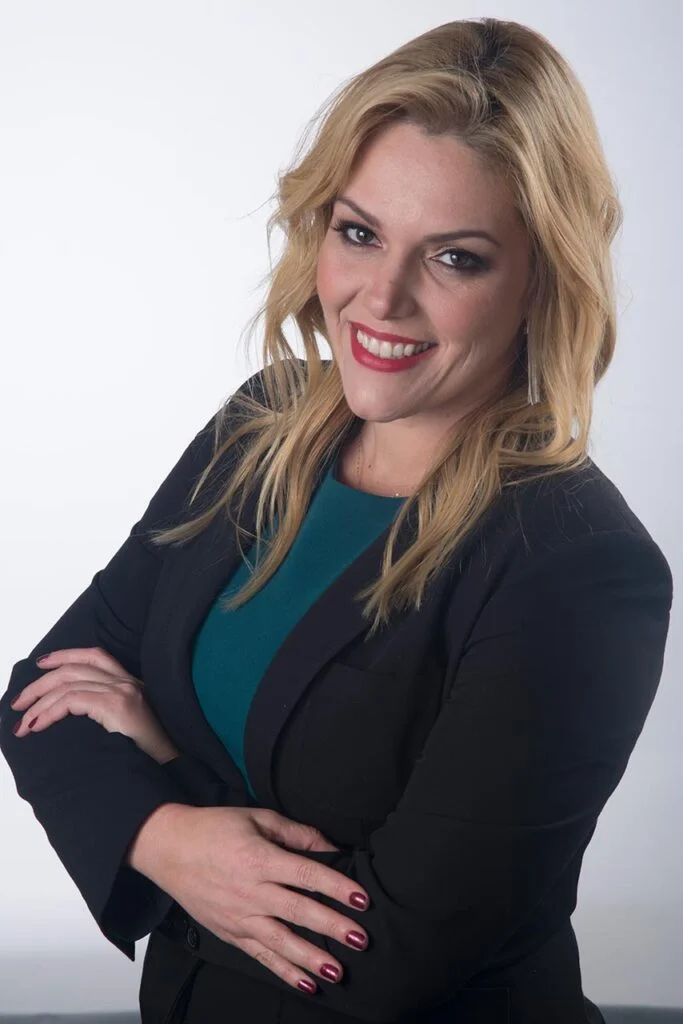 retrato profissional de mulher loira, jovem, sorrindo, vestindo blazer preto e blusinha verde escuro, braços cruzados, ambiente claro
